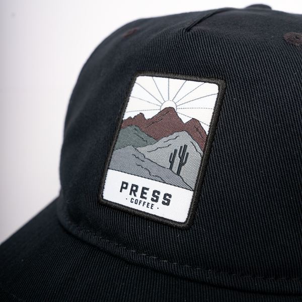 A close up view of a Press Coffee hat featuring a patch with a cactus and desert mountains.