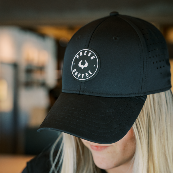 A sleek black hat featuring a circular Press Coffee logo is worn by a blonde woman.