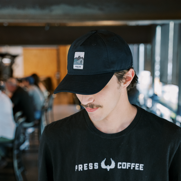 A man with a mustache wearing a Press Coffee dad hat featuring a hat with patch that includes cactus, a desert mountain range, and a beaming sun.