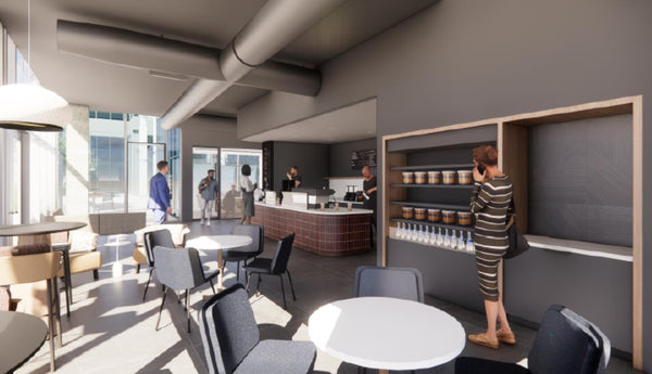 A modern interior cafe or breakroom space with round white tables, gray chairs, a counter, shelving, and several people visible in the space.