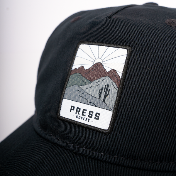 A close up view of a Press Coffee hat featuring a patch with a cactus and desert mountains. 