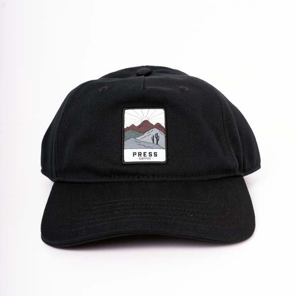 A full view of a Press Coffee dad hat with a patch that features a cactus and desert mountain range. 