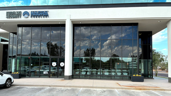 A modern commercial building facade with large black-framed glass windows, a white exterior wall, and visible Kudaek Security and Nagra Security signage above the entrance. A small white vehicle is partially visible at the lower left, and the scene is photographed outdoors under a blue sky with cloud reflections in the glass.