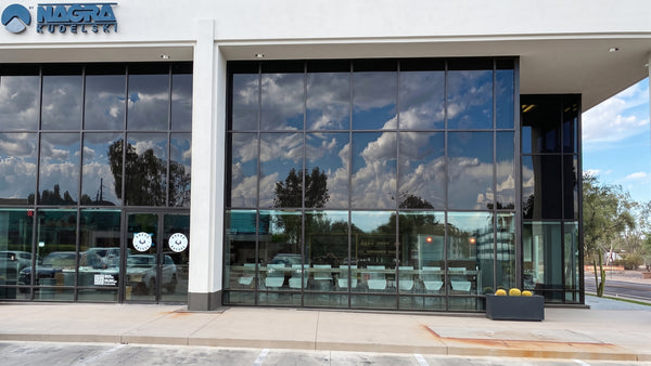 The exterior of a modern building featuring large black-framed glass windows reflecting a cloudy blue sky. The white facade has a blue and white 'NAGTA ASSOCIATES' logo on the upper left, and a concrete sidewalk runs along the front.