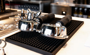 Two polished chrome portafilter baskets with black handles resting on a black textured rubber mat on a white countertop.