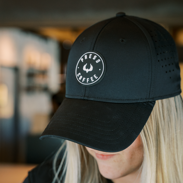 A sleek black hat featuring a circular Press Coffee logo is worn by a blonde woman. 