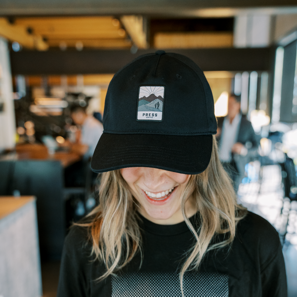 A smiling young woman wears a Press Coffee hat featuring cactus and a desert mountain range. 