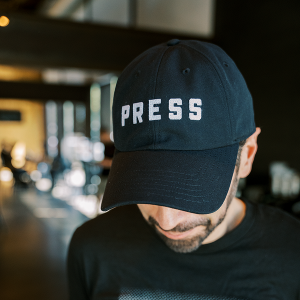 A man with a beard wearing a PRESS dad hat. 