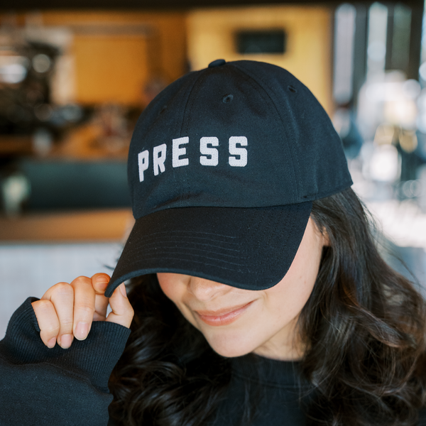 A woman with dark hair wearing a PRESS hat. 