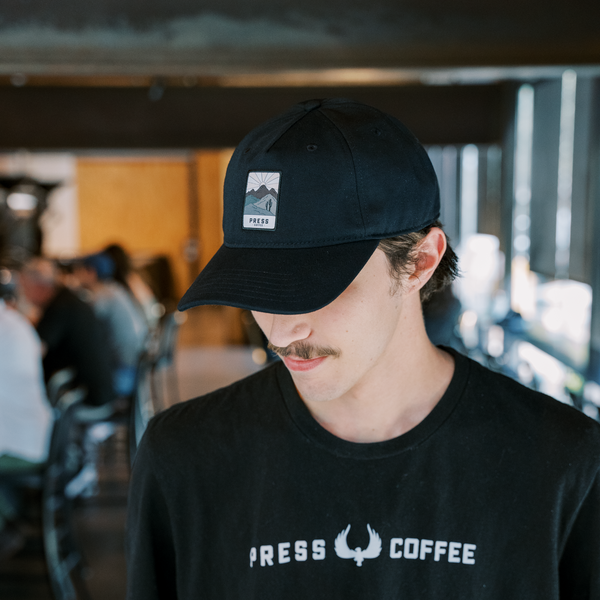 A man with a mustache wearing a Press Coffee dad hat featuring a hat with patch that includes cactus, a desert mountain range, and a beaming sun. 